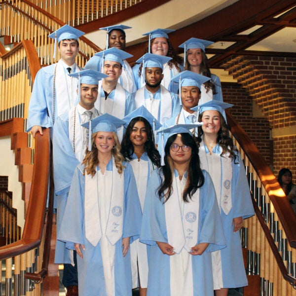 12 Radiologic Science graduates wearing cap and gowns standing on the stairs in the lobby of the MBRB building.