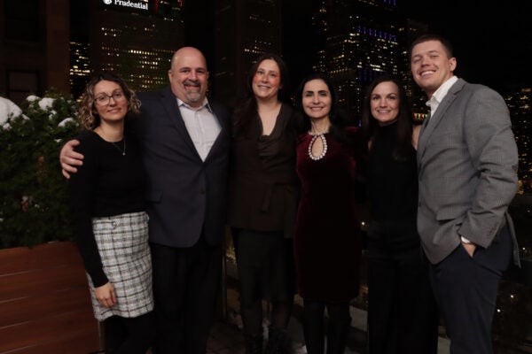 Six adults standing close together and smiling for a group photo at night on an outdoor rooftop patio. Chicago skyscrapers with illuminated windows form the background, and snow-dusted greenery appears on the left. The group is dressed in semi-formal attire, with arms around one another.