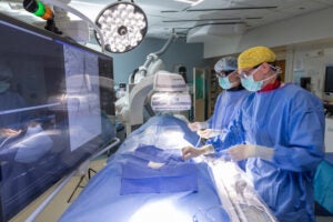 Two medical professionals in blue scrubs and face masks stand beside a patient lying on a hospital bed. One is adjusting equipment while the other monitors a screen. The setting is a clean, modern clinical environment, emphasizing attentive patient care.