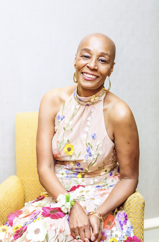 Photo of Wendy Brewster smiling and sitting in a yellow chair. She is wearing a dress embroidered with flowers.